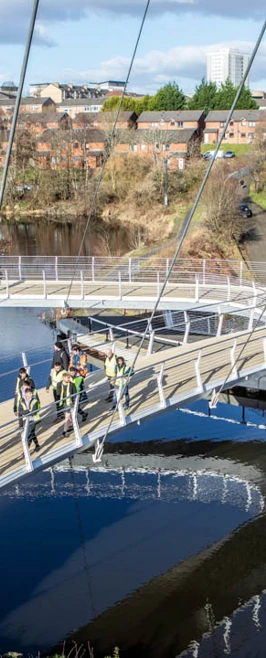 Overhead view of students and lecturers walking across the curved paths of the Stockingfield Bridge, with the canal reflecting the structure and surrounding landscape. Overhead view of students and lecturers walking across the curved paths of the Stockingfield Bridge, with the canal reflecting the structure and surrounding landscape.